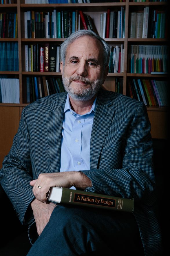 Alex Aleinikoff sits, smiling into the camera, holding a book on his lap