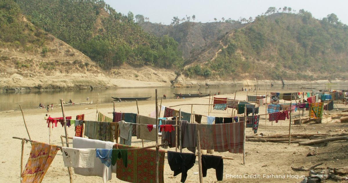 Colourful laundry dries on frames made of tall branches in front of a body of water in teh Chittagong Hill Tracts. There are canoes of the water, with people in, and small children play on the bank. In the background, tree covered hills.