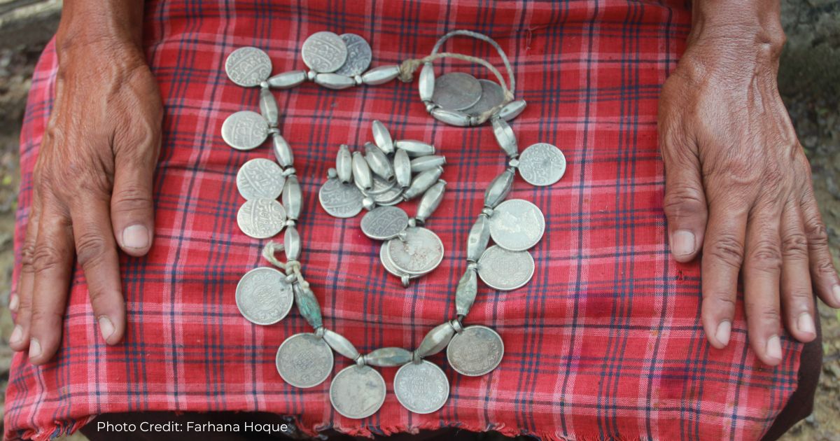 A coin garland is displayed in the lap of a Marma woman. We see her hands rest in her lap on either side of the pieces. 