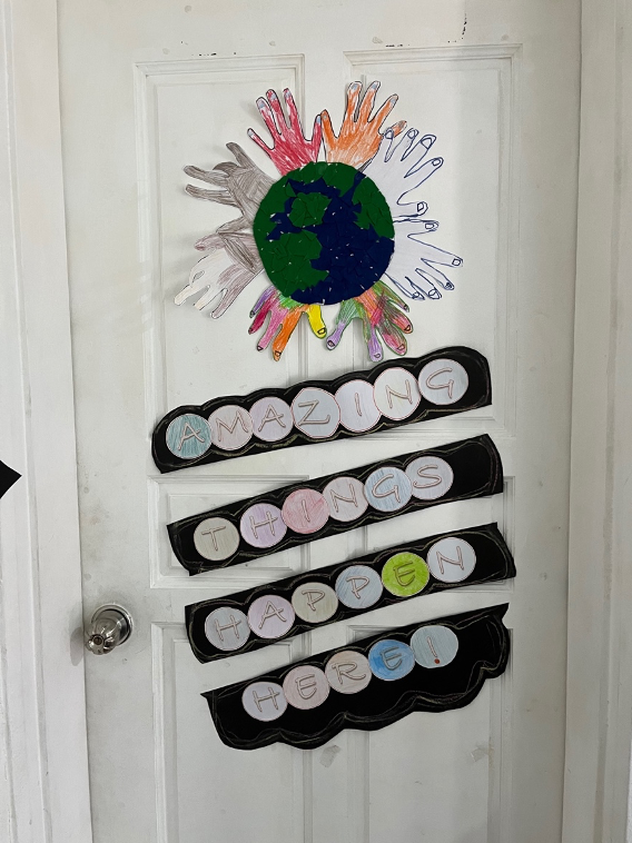 Children's schoolwork glued to a white door featuring a world with white and coloured hands around it and a colourful phrase with letters on a black background saying 'Amazing things happen here!'.