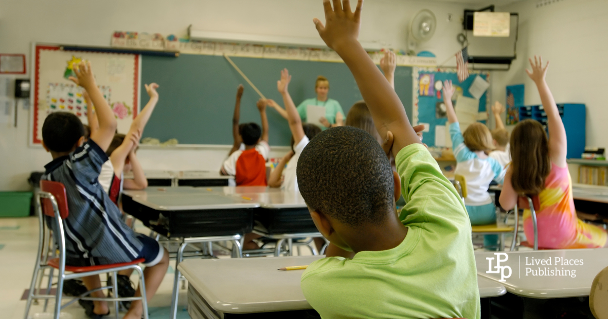 Image of an elementary school classroom from the back of the room with students raising their hands