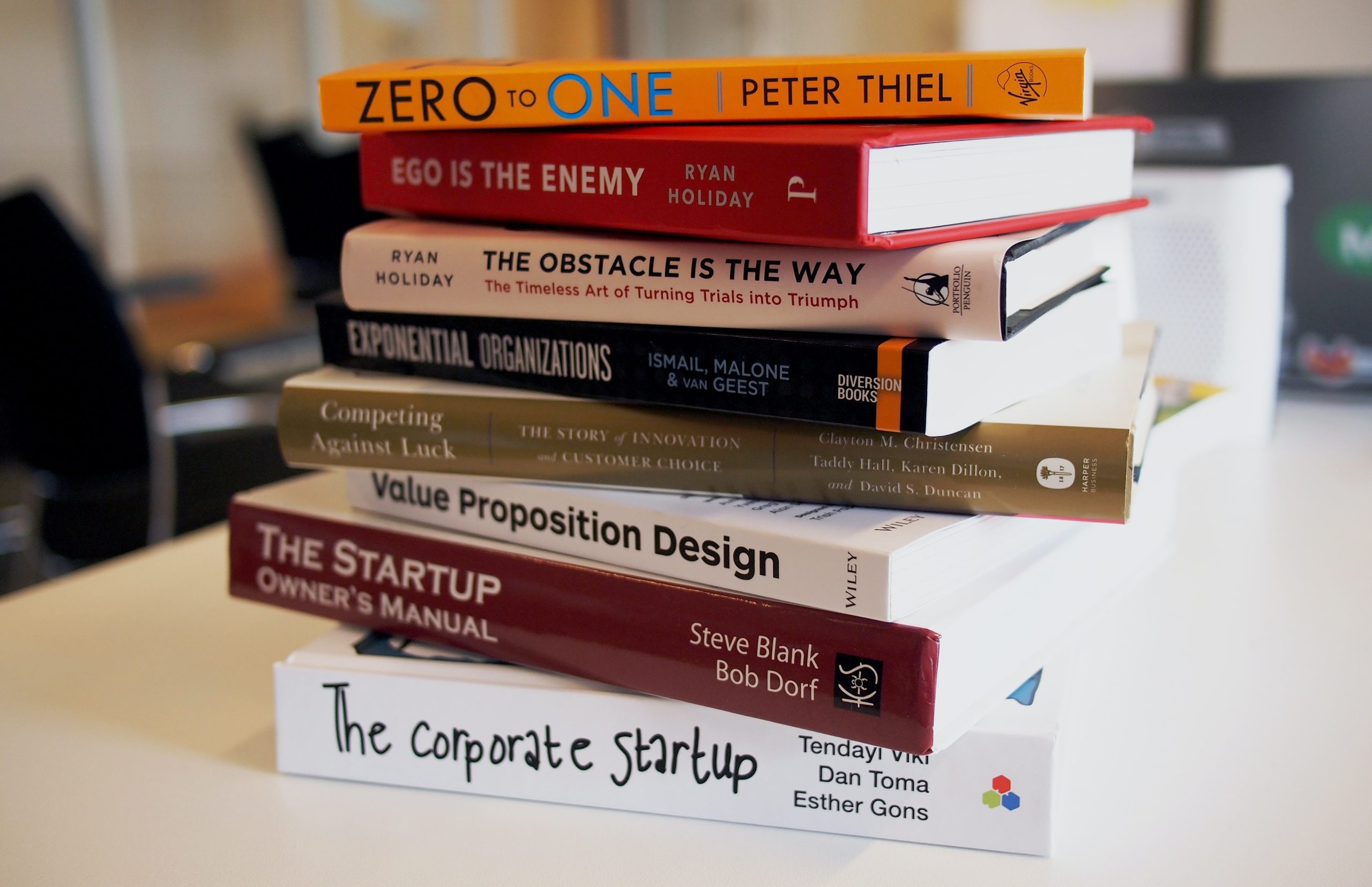 On a white desk in an office lies a higgledy-piggledy stack of book, colorful spines visible. They are all popular entrepreneurship titles, such as “ Zero to One” by Peter Thiel, and “The corporate startup” by Tendayi Viki, Dan Toma and Esther Gons.