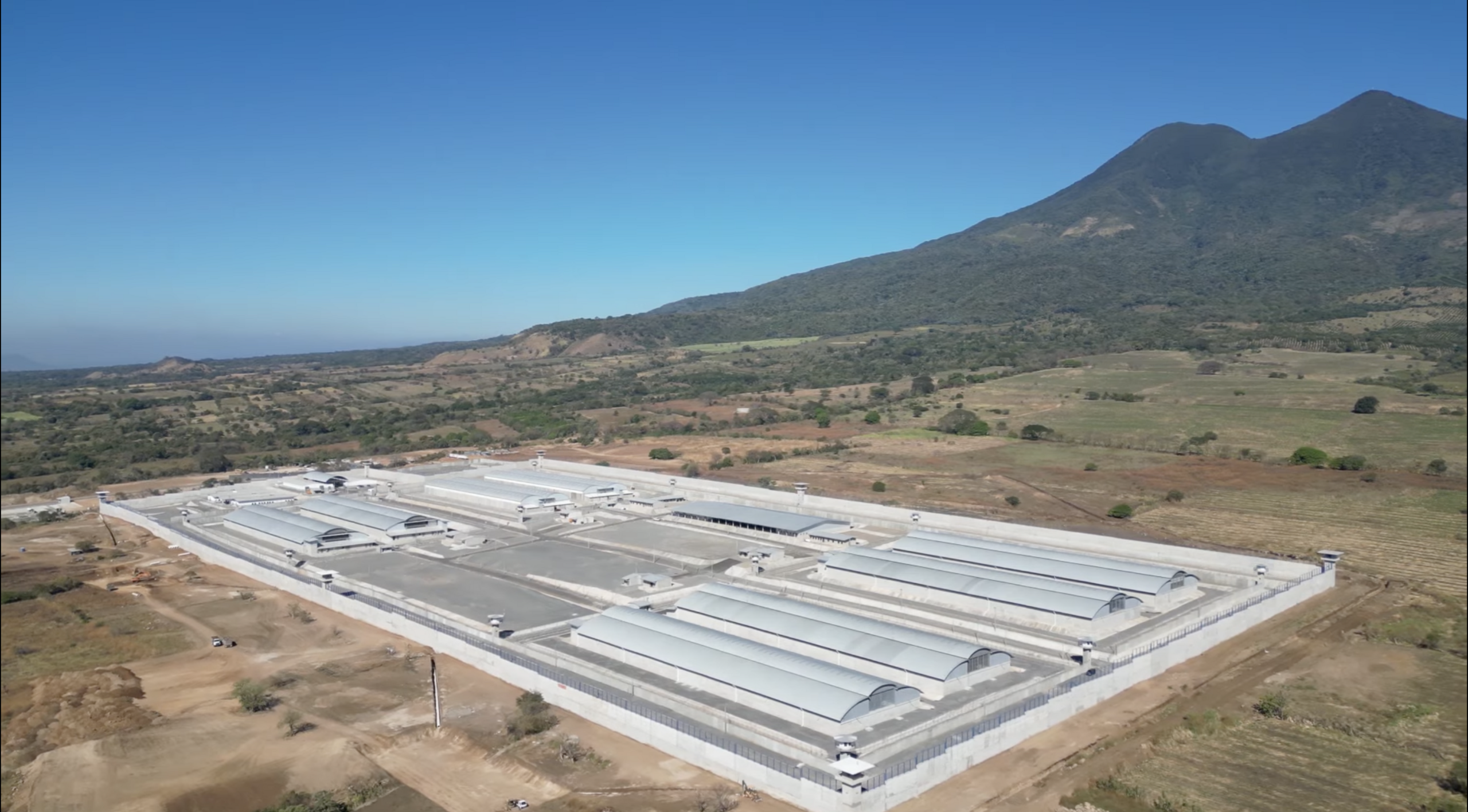 Aerial view of CECOT, the maximum security prison in El Salvador – a large, isolated industrial building with a mountain and sky in the background.