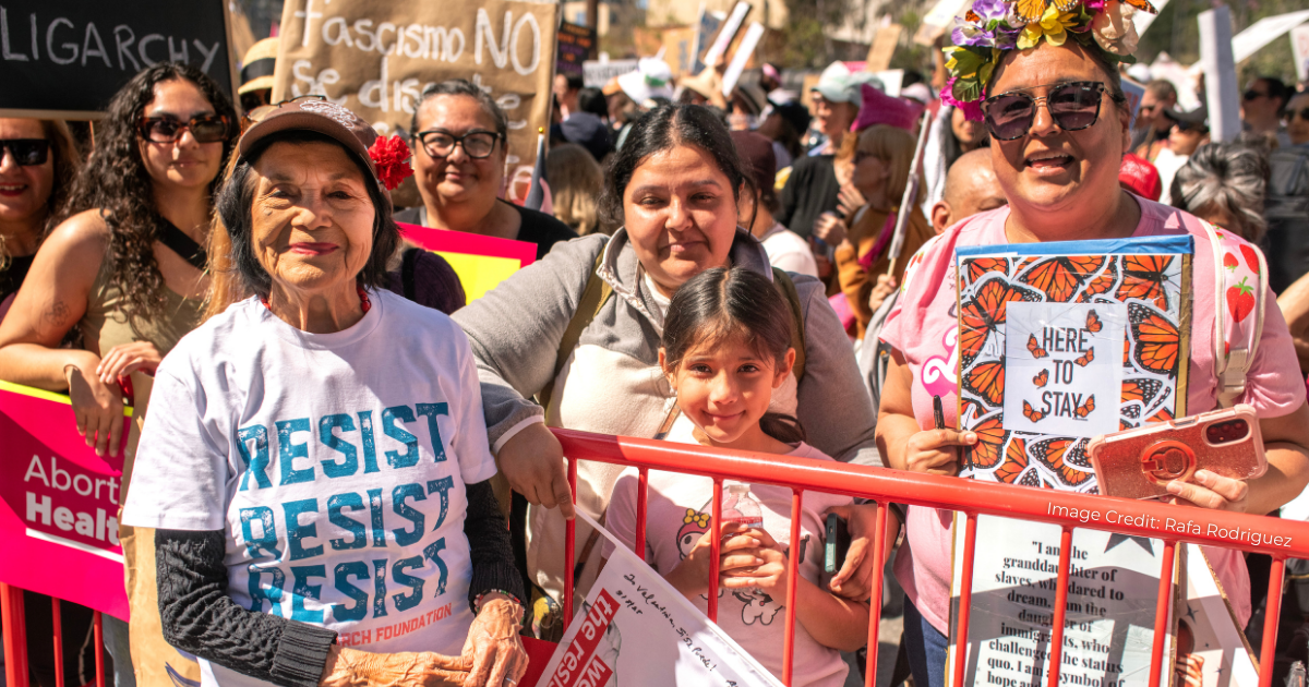Activist Dolores Huerta is in the foreground at left, wearing a shirt that reads "RESIST RESIST RESIST," at a demonstration for migrant rights photographed with other activists, one of whom is holding a placard that reads, "HERE TO STAY."