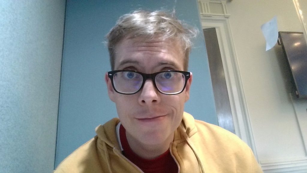 Headshot of man with blonde hair wearing glasses and a yellow hoodie