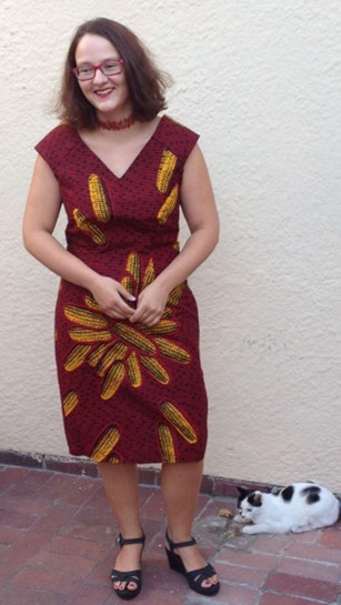 A full-length photograph of a smiling woman with red hair in a maroon sheath dress with a pattern of corn cobs.