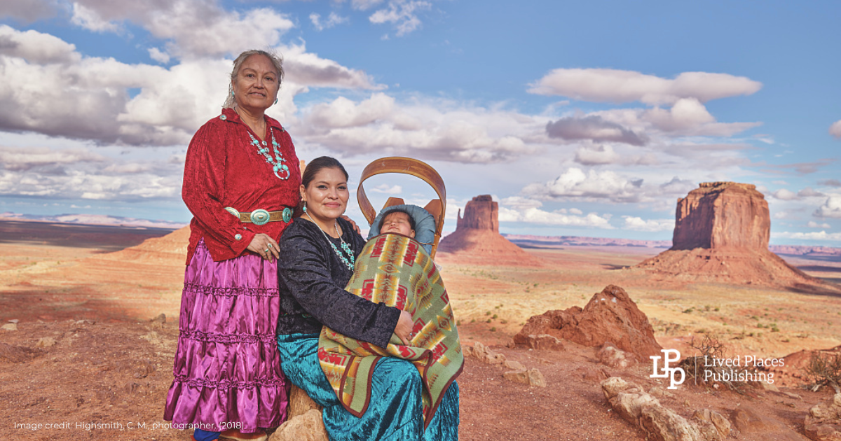 Three generations of Navajo women, a grandmother standing and a mother seated holding an infant child – all in traditional dress outdoors in Monument Valley Navajo Tribal Park.