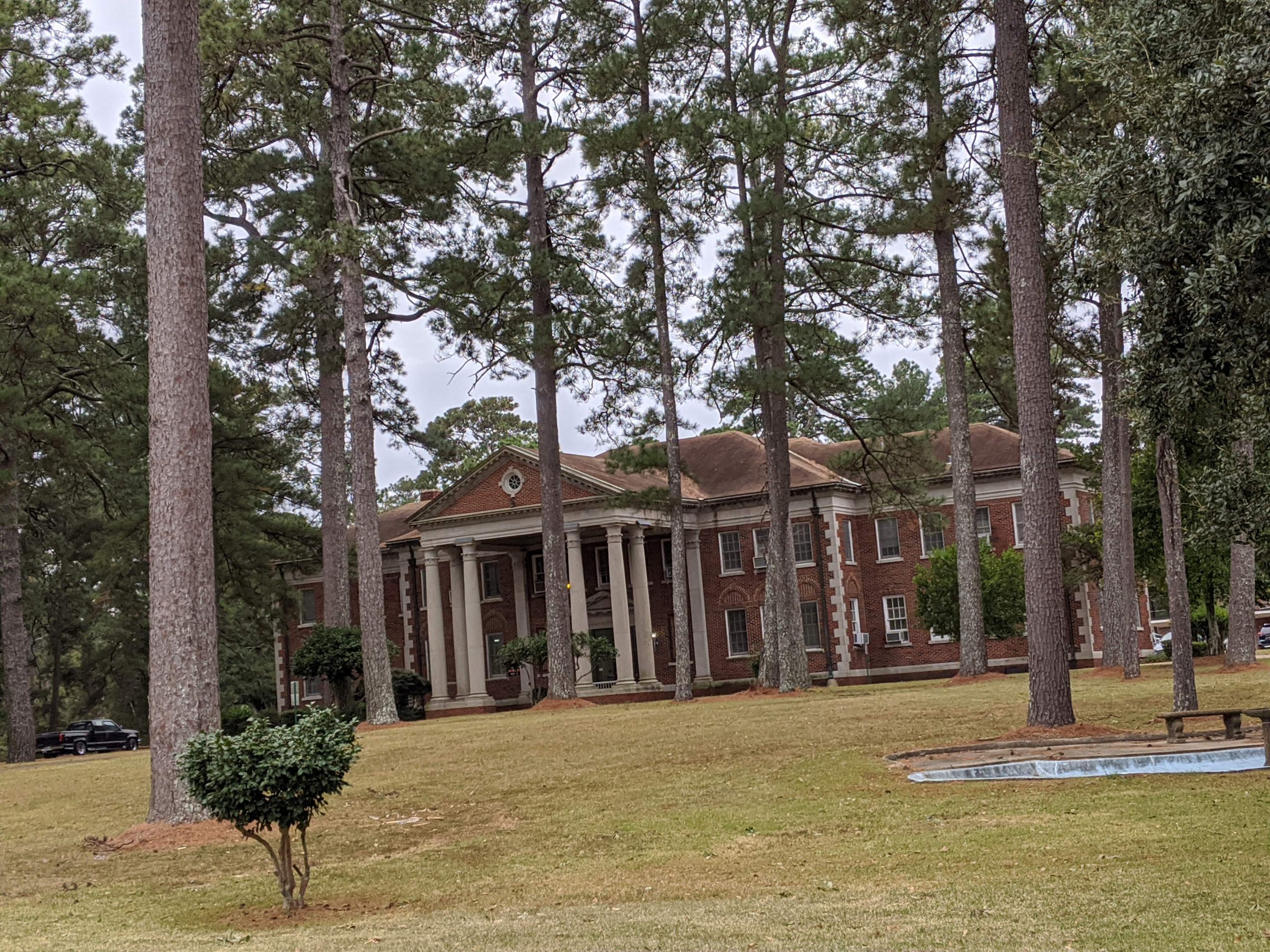 A two-story red brick building with white pillars surrounded by tall pine trees and a green lawn.
