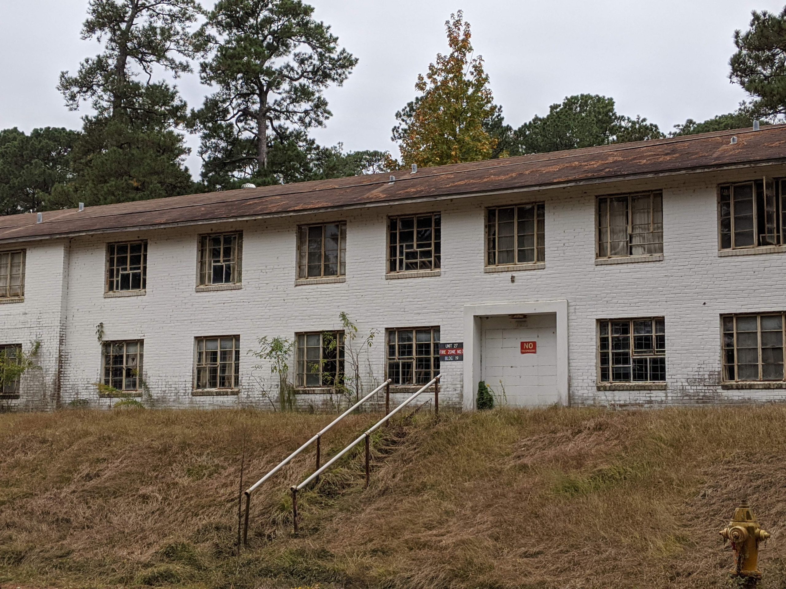 A two-story white brick building in disrepair with broken windows and surrounded by overgrown brush and grass. A flight of cement steps and a metal railing lead to the front door.