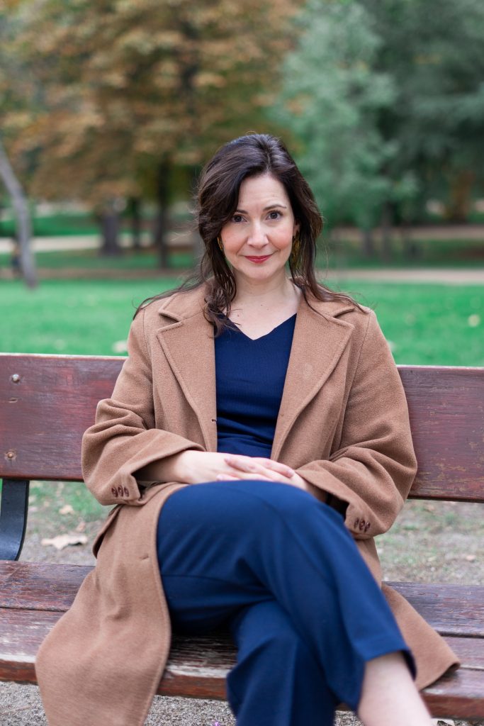 A picture of a woman seated on a park bench wearing a dark blue shirt and pants with a brown overcoat. Dr. Angy Cohen is a Ramón y Cajal Researcher at the Spanish National Research Council (Madrid, Spain) and Adjunct Professor at University of Calgary (Canada).