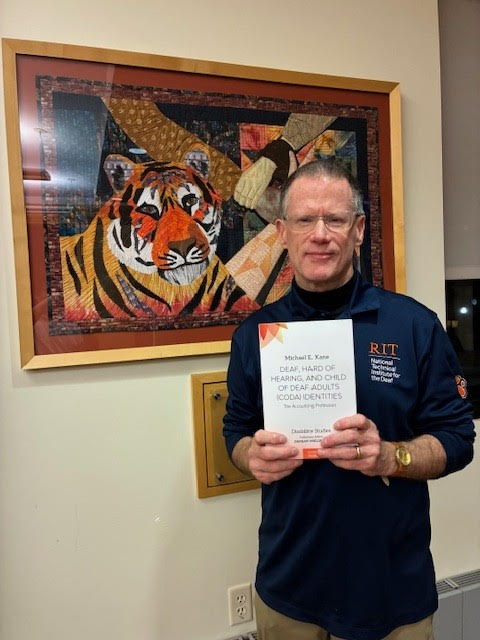 Michael Kane holding "Deaf, Hard of Hearing, and Child of Deaf Adults (CODA) Identities" in front of a painting of a Tiger.