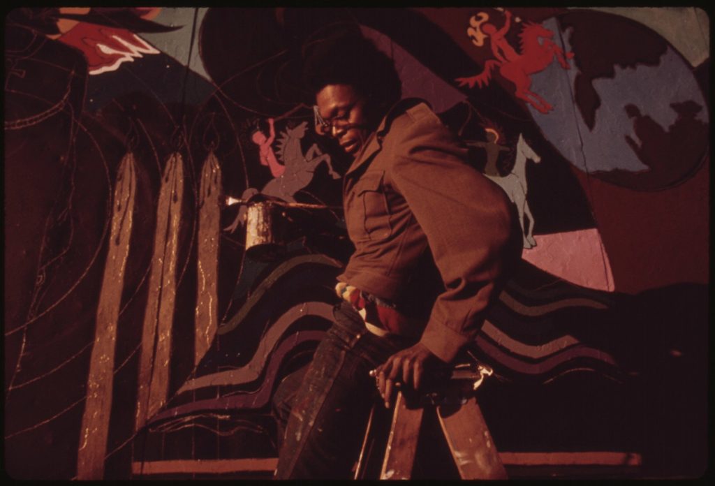 Black man painting a multicolored wall with a brown jacket on while sitting on a wooden stool. 